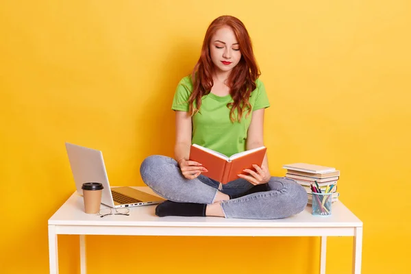 红头发浓密的女性 身穿绿色T恤和牛仔裤 手里拿着书本看书 学生们双腿交叉坐在桌子上 女士身边围着笔记本电脑 — 图库照片