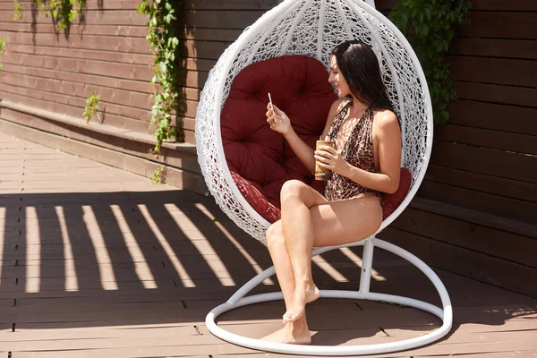 Üzerinde leopar desenli mayo olan esmer kadın beyaz şık bir sandalyede oturuyor ve video görüşmesi yapıyor ya da selfie çekiyor, kokteylden keyif alıyor..