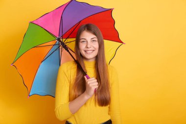 Çok renkli bir şemsiye tutan güzel bir kadın ve doğrudan kameraya bakıyor, sarı duvara karşı poz veriyor, günlük kıyafetler giyiyor, uzun saçlı bir kadın..