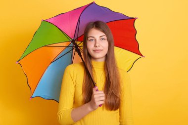 Çok renkli şemsiyelerin altında duran, doğrudan kameraya bakan, sarı kazak giyen, uzun saçlı, sakince yüz ifadesi olan çekici genç bir kadın..