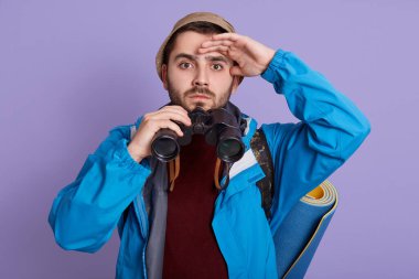 Dürbün tutan, eli alnına yakın, mesafeye bakan genç adam ciddi görünüyor, günlük kıyafetler ve şapka giyiyor, leylak duvarın önünde duruyor..