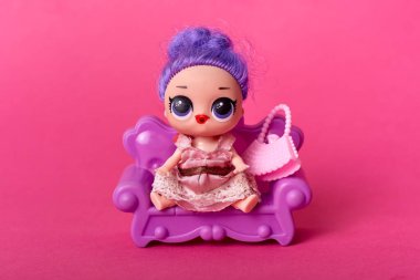 Mor saçlı, büyük gözlü, pembe elbiseli modern çocuk oyuncağı, pembe arka planda küçük koltukta oturan büyük gözlü bebek..
