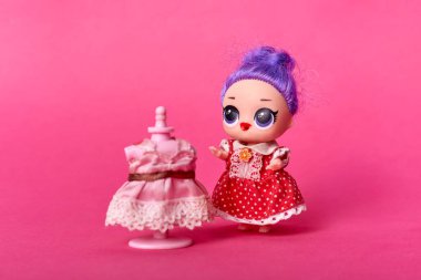 Mor saçlı, büyük gözlü, kırmızı benekli elbise giyen, güzel pembe elbiseli, pembe arka planda izole bir mankenin yanında poz veren sevimli bir bebek..