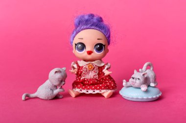 Etrafı iki oyuncak kediyle çevrili şirin bir oyuncak bebek mor saçlı, büyük gözlü, benekli elbise giyen, gül arka planında izole edilmiş, kızlar için oyuncaklar..