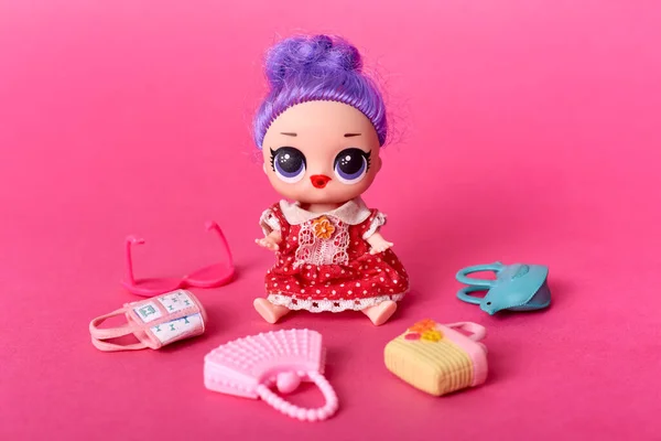 Toys for girls, doll with big eyes and lilac hair, purple headed tiny doll surrounded with small toy bags and glasses, isolated over rosy background, child choose bag for toy doll..