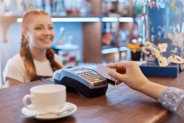 Müşteri cep telefonuyla kahve dükkanında elektronik okuyucu üzerinden ödeme yapıyor. Yüzsüz müşteri kartı tutuyor. Tezgahın üzerinde gülümseyen kızıl saçlı garson..