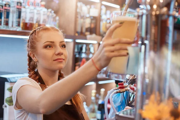 Female bartender tapping beer in bar, red haired barmen posing near counter, has pigtails, standing with showcase on background, looking at glass with concentrated look.
