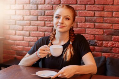 Kızıl saçlı güzel bir kadın elinde beyaz seramik bardakla kafede oturmuş sıcak kahvenin tadını çıkarıyor tuğla duvara karşı poz veriyor ve kameraya bakıyor..