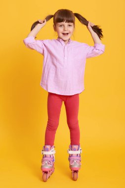 Küçük kız sarı arka planda paten kayıyor, gömlek ve tayt giyiyor, saçına dokunuyor, doğrudan kameraya bakıyor, tatlı bayan çocuk eğleniyor..