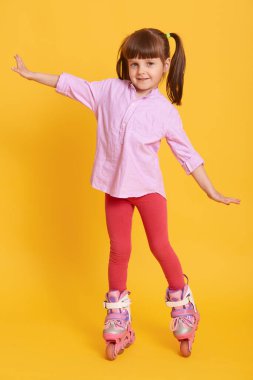 Hoş görünümlü bir çocuk patenlerle el kaldırıyor, iki at kuyruğu takıyor, sarı arka planda tek başına duruyor, eğleniyor ve kameraya bakıyor..