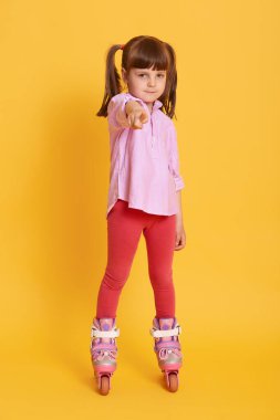 Paten giyen, ön parmağıyla kameraya bakan ve işaret eden çekici kadın çocuk, gömlek ve tozluk giyen çocuk, sarı arka planda poz veren iki at kuyruklu..