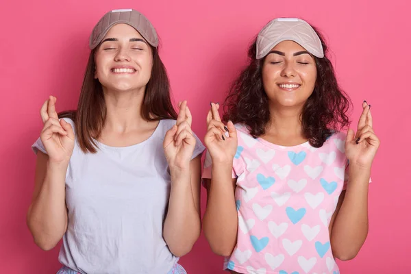 Two pretty women in pajamas and blind fold praying with crossed fingers, keep eyes closed, standing against pink wall, ladies with toothy smiles, wearing pajamas.