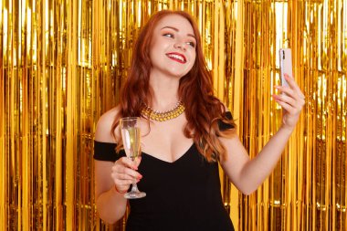 Mutlu bir gülümseme ile altın rengi süslemelere karşı poz vermek ve modern akıllı telefondan selfie çekmek, siyah elbiseli bir kız, elinde şarap kadehi tutan bir kadın, önemli bir olayı kutluyor..