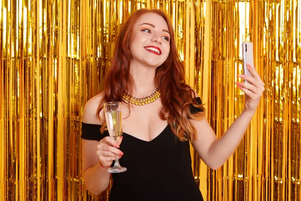 Mutlu bir gülümseme ile altın rengi süslemelere karşı poz vermek ve modern akıllı telefondan selfie çekmek, siyah elbiseli bir kız, elinde şarap kadehi tutan bir kadın, önemli bir olayı kutluyor..