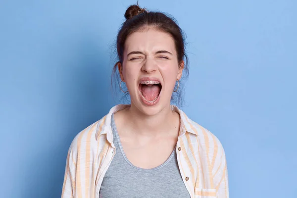 Young emotional angry woman screaming isolated over blue background,keeping eyes closed, looks sad, wearing white shirt and grey t -shirt, brunette lady with knot yelling angrily.