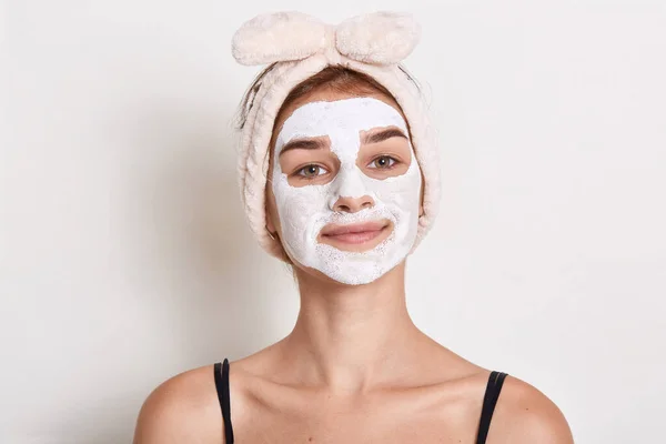 Adorable young female with white facial beauty mask looks directly at camera, girl with hair band doing beauty procedure, posing against white wall in sleeveless t shirt.