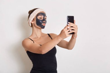 Yüzünde siyah kozmetik maskeli bir kızın portresi modern akıllı telefonu elinde tutuyor ve cihaz ekranına bakıyor, beyaz arkaplanda izole edilmiş selfie yapıyor..