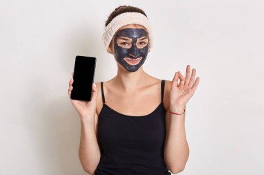 Saç bandı ve tişört giyen, gülümseyen, telefonu boş ekranla tutan, ince parmaklar gösteren, beyaz arka planda soyutlanmış poz veren çekici bir kadın..