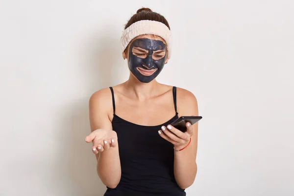 携帯電話を使用して黒い顔の化粧品マスクを持つ若い女性 デバイス画面を直接見て 手のひらを広げて 驚きと困惑顔の表情を持っています — ストック写真