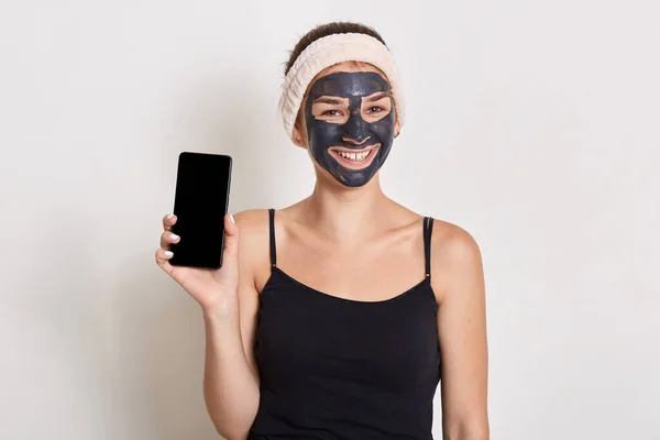 顔に黒の化粧品マスクを持ついくつかの笑顔の女の子 空白の画面で携帯電話を保持 広告のためのスペースをコピー 女性は自宅で美容手順を行う — ストック写真