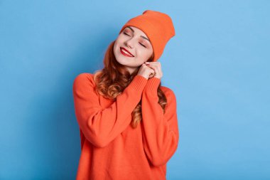 Güzel bir kadın dişlek, elleri kulağın yanında, turuncu şapka ve kazak giyiyor, yeni kıyafetlerle sıcacık, mavi duvarda izole edilmiş, hayaller kuruyor, hoş bir şeyler hayal ediyor..