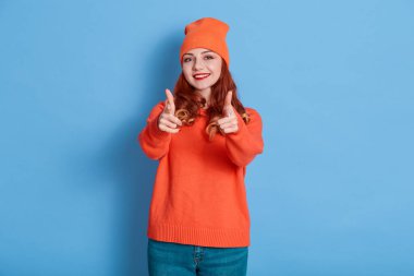 Heyecanlı kızıl saçlı kadın, mavi arka planda izole edilmiş kamerayı işaret ediyor. Kadın dişlek gülümsüyor. Beyaz dişli, kızıl saçlı kız kameraya bakıyor..