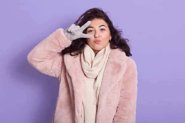 若い美しいブルネットの女性はピンクの冬の毛皮のコートを着て 幸せな顔で立って カメラを見て 彼女の目の前で勝利サインをして 唇を丸めて キスジェスチャーを作る — ストック写真