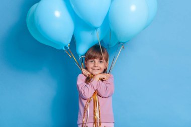 Mavi arka planda helyum balonları olan mutlu çocuk, pembe kazak giyen kadın çocuk, kameraya bakıyor, doğum gününü kutluyor, çocuk partisine gidiyor, pozitif duygularını ifade ediyor..