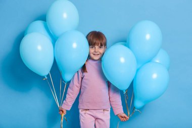 Mutlu bir çocuk, mutlu bir doğum günü partisinde pembe kazak ve pantolon giyer, kameraya büyüleyici bir gülümsemeyle bakar, her iki elinde de mavi balon demetleri tutar..