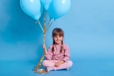 Siyah saçlı, saçları örgülü, ayakları çapraz yerde oturan, kameraya bakan, helyum balonları tutan, mavi duvara karşı günlük kıyafetler giyen, doğum günü partisi veren bir kız çocuğu..