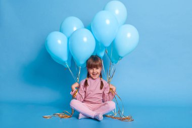 Sevimli küçük bir kız elinde balonlarla yerde oturuyor, kameraya bakıyor ve gülüyor, günlük kıyafetler giyiyor, keyfi yerinde, yerde oturuyor ve mavi duvara karşı bacak bacak bacak üstüne atıyor..