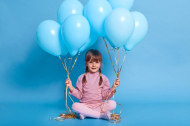 Siyah saçlı, iki örgülü bir kız çocuğu kameraya bakıp sevimli bir gülümseme takınıyor. Elinde balonlar, pembe elbiseli sevimli bir çocuk yerde bacak bacak bacak üstüne atmış bir şekilde oturuyor ve doğum gününü kutluyor..