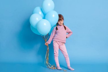 Mavi kalçalı, aşağı bakan, mavi duvara poz veren şirin kız anaokulu öğrencisinin portresi, iki örgülü, helyum balonlu, reklam için kopyalama alanı olan.