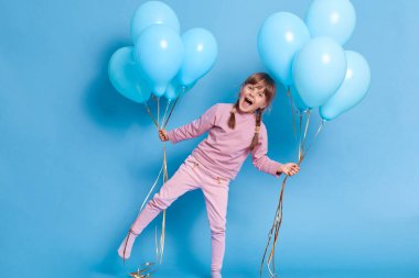 Tek bacağı üzerinde duran mutlu, ağzı açık, siyah saçlı ve örgülü bir kız elinde helyum balonları, mavi arka planda izole bir şekilde poz veriyor, doğum günü partisinde eğleniyor..
