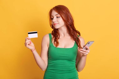 El ele tutuşan güzel kızıl saçlı bir kızın portresi banka kartı ve cep telefonu, kablosuz alışverişi, sarı arka planda izole edilmiş şeyleri internetten alan kızıl kız..