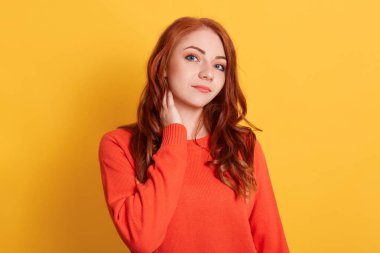 Güzel kızıl kız elini boynuna koyuyor, morali yüksek, sarı arka planda poz veriyor, sakin bir ifadeyle kameraya bakıyor, günlük turuncu kazak giyiyor..