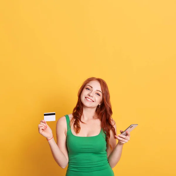 Happy beautiful lady with red hair and charming smile, being in good mood, having unlimited credit card, holding phone in hands, wearing green dress against yellow background. Copy space for promo.