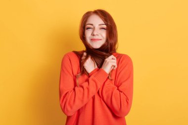 Çekici, çekici kızıl saçlı, parlak sarı arka planda izole edilmiş saçları olan, turuncu kazak giyen güzel ve çekici bir kızın yakın plan portresi tatmin olmuş görünüyor..