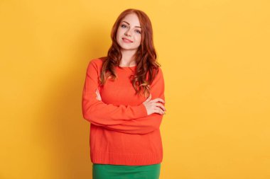 Kafkas görünümlü genç bir kadının portresi günlük giysiler giymiş, kolları çaprazlanmış boş bir duvara poz veriyor, mutlu hissediyor, gülümsüyor, turuncu kazaklı kızıl saçlı kadın..