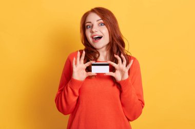 Mutlu, heyecanlı, genç bir kadın elinde kredi kartıyla kameraya bakarken ağzı açık, kırmızı saçlı, sarı duvarda turuncu kazak giyen bir kadın..