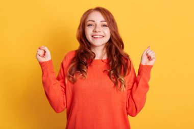 Aşırı neşeli kızıl saçlı genç bayan, iki yumruğunu sıkıyor, başarıyı kutluyor ve sevinçle haykırıyor, yaşasın jestler yapıyor, başarılı anlaşmalar yapıyor, kameraya bakıyor, günlük kazak giyiyor..