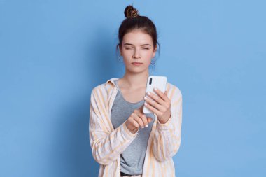 Esmer kadın saçını tarıyor, modern cep telefonu tutuyor, mavi arka planda izole edilmiş günlük gömlek giyiyor, esmer kadın mesajlar yazıyor, cihazın ekranına bakıyor..