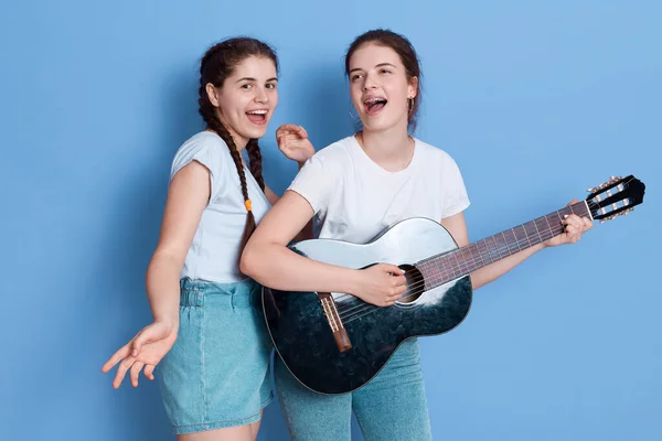 2つの美しい豪華な愛らしい女性は 青い背景の歌と演奏ギター 白いTシャツとジーンズを身に着けている女の子 ブレース付きの女性は楽器を保持しています — ストック写真