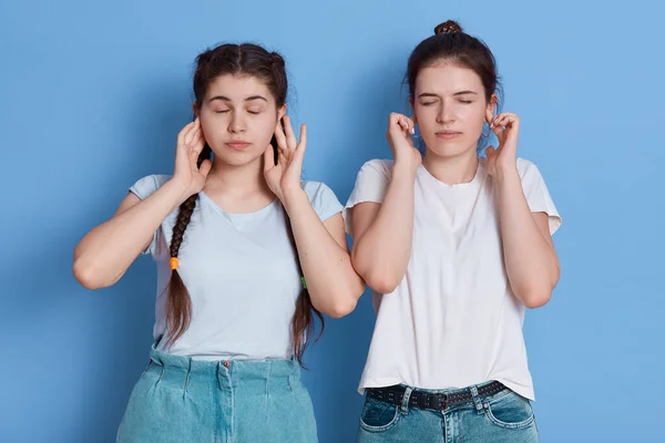 十代の友人不満と手で耳をカバー 非常に大きな音楽を聞くことができません 目を閉じてください ブルネットの女性は白いカジュアルなTシャツを着て 青の背景に孤立立って — ストック写真