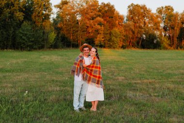 İki insan, gün batımında huzurlu bir çimenlikte birlikte gülümseyen renkli bir battaniyeye sarılı sıcak bir anı paylaşır..