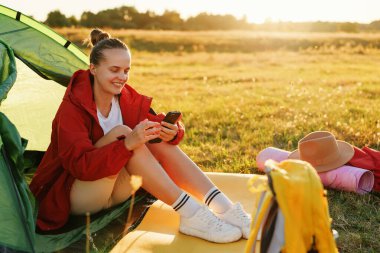 Neşeli genç bir kadın çadırın yanında oturuyor, akıllı telefonuna gülümsüyor ve sıcak güneş ışığının tadını çıkarıyor..