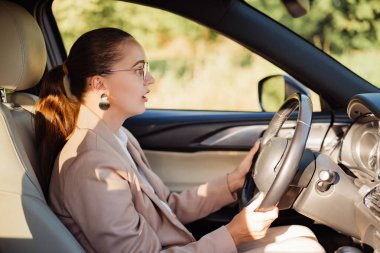At kuyruğu ve gözlüklü endişeli bir kadın araba sürerken önüne bakar, ağzı şaşkınlık ve endişe içinde hafifçe açılır..