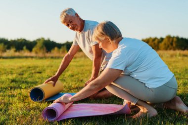 Barışçıl bir alanda, bir çift yoga minderlerini çimlerin üzerine serer, güneş arkalarında batarken birlikte bir seansa hazırlanırlar, sakin bir atmosfer yaratırlar..