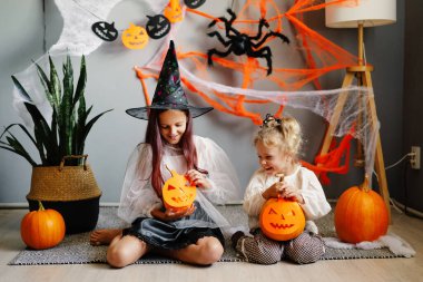 Cadılar Bayramı için dekore edilmiş bir odada gülümseyen ve balkabağı tutan cadı ve peri kostümlü iki genç kızın samimi bir fotoğrafı..