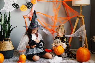 Cadılar Bayramı 'nda süslü kostümlü iki kız kardeşin, yerde balkabağı ve dekore edilmiş evlerinde örümcekle otururken gülümserken çekilmiş samimi bir fotoğrafı..